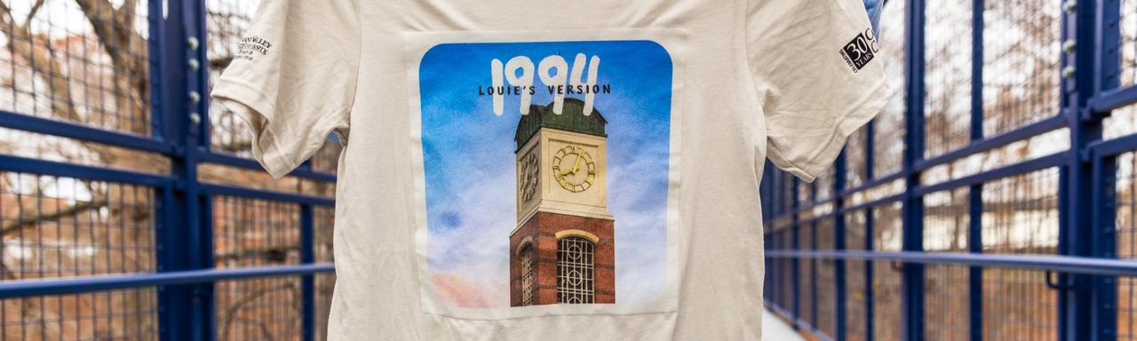 A student holding a beige t-shirt featuring a clock tower and text that reads "1994. Louie's version."